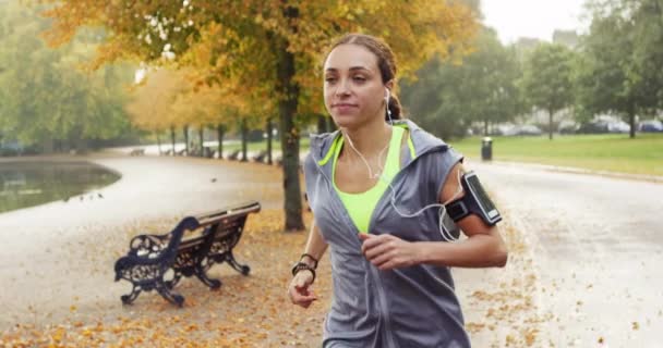 A young woman using her cellphone to listen to music through earphones while jogging through the park. A young woman exercising and running through a garden listening to music in slow motion. — Stockvideo