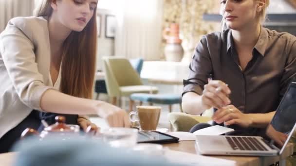 Two businesswomen working together brainstorming during a meeting in a cafe and drinking coffee. Businesswomen collaborate to work on documents, a digital tablet and a laptop in a cafe. – stockvideo