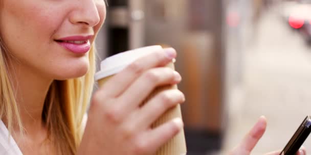 Молода бізнес-леді п'є каву, використовуючи свій мобільний телефон. Молода бізнес-леді насолоджується чашкою чаю і використовує свій мобільний телефон під час перебування у місті. Молода жінка використовує свій смартфон зовні — стокове відео
