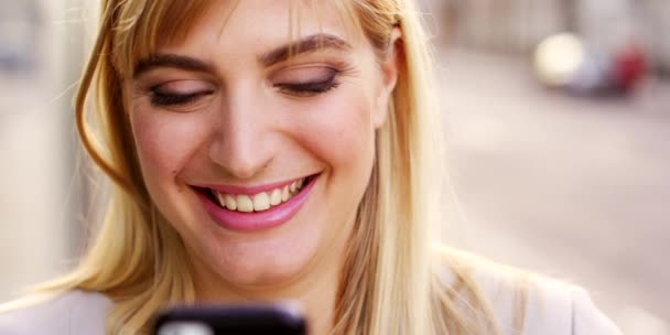 A businesswoman using her smartphone while walking to work. Cheerful businesswoman laughing while reading texts on her cellphone outside in the city. Businesswoman using an app on her smartphone — Stockvideo