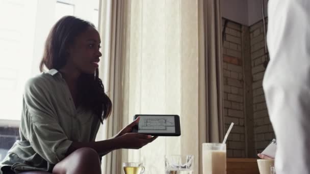 A diverse group of architects looking at blueprints on a wireless tablet while sitting in a cafe. A group of businesspeople planning, brainstorming and talking during a meeting. — Vídeo de stock