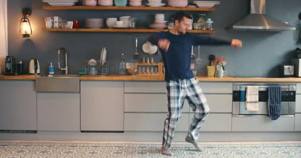 A young man using a phone while dancing in the kitchen at home. Carefree playful young man having fun, dancing in his kitchen to music from his cellphone. Cheerful young man dancing in his pyjamas — Video