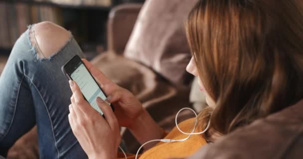 Πίσω από μια γυναίκα που κάθεται σε έναν καναπέ χρησιμοποιώντας διαδικτυακές εφαρμογές στο κινητό της, ακούγοντας μουσική. Μια γυναίκα που χαλαρώνει στο σπίτι στέλνοντας μήνυμα στο κινητό της. Γυναίκα που ακούει μουσική στο τηλέφωνό της — Αρχείο Βίντεο