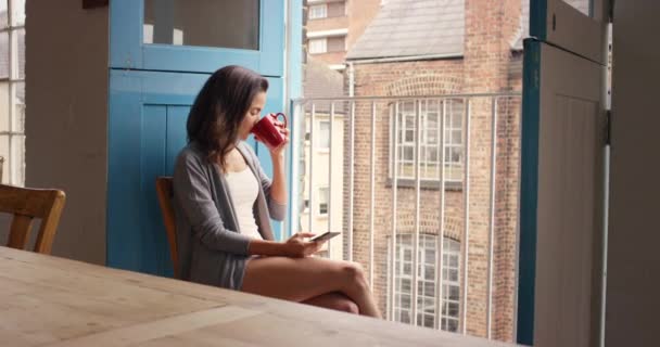 Uma jovem mulher usando um telefone enquanto bebe café em casa. Conteúdo jovem mulher sentada em sua cozinha desfrutando de uma xícara de café, lendo uma mensagem de texto em seu celular. Jovem relaxante em casa — Vídeo de Stock