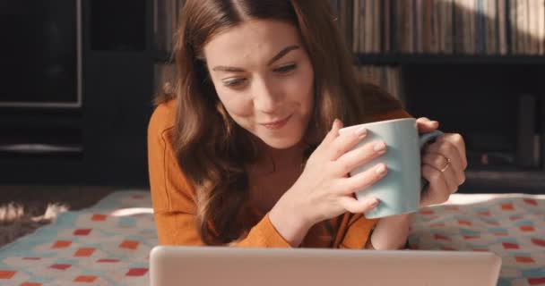 Egy fiatal, aki laptopot használ, miközben otthon kávézik. Egy boldog fiatal nő fekszik, kávézik és otthon használja a laptopját. Elégedett fiatal nő feküdt a padlón teát iszik — Stock videók