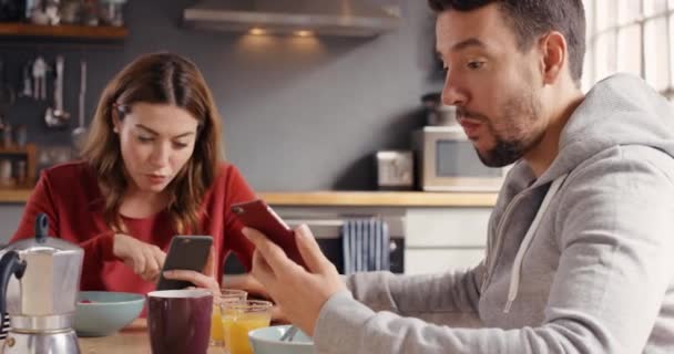 A young couple using their phones while having lunch together at home. A happy young man showing his girlfriend his smartphone while eating breakfast together in the kitchen. A couple eating together – Stock-video
