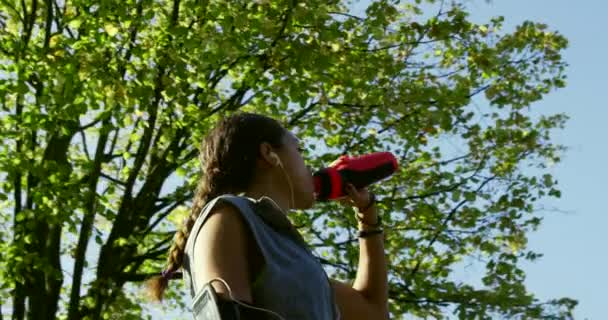 Jeune femme prenant une pause de jogging dans le parc pour boire de l'eau. Ajuster jeune femme à écouter de la musique à l'aide d'écouteurs prendre une pause de son entraînement dans le jardin pour boire de l'eau d'une bouteille — Video