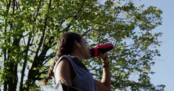 Mujer joven tomando un descanso de su entrenamiento para beber agua mientras escucha música en el parque. Una joven escuchando música a través de sus auriculares mientras bebe agua de una botella en un jardín — Vídeo de stock