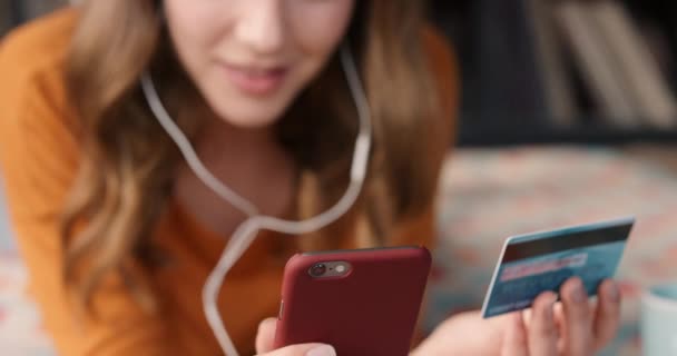 Young caucasian woman happy smiling relaxing using phone holding credit card hand listen music at home. Smiling woman listening to music paying for online shopping with her debit card on her phone — Stockvideo