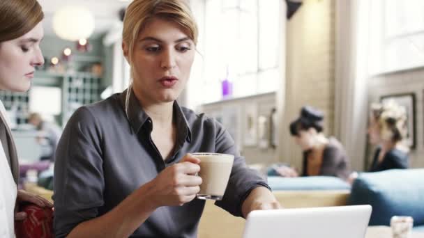 Dos empresarias colaboran en una oficina, hablando mientras usan un portátil. Una joven jefa en una reunión con su colega, tomando una taza de café mientras planean un proyecto de marketing juntos. — Vídeo de stock