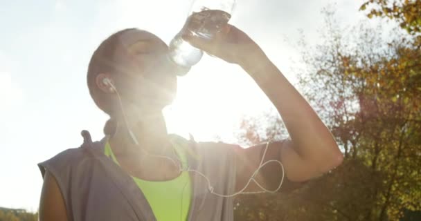 Una joven bebiendo agua de una botella y escuchando música mientras se toma un descanso durante su entrenamiento. Una joven haciendo ejercicio en el parque tomando un descanso para beber agua. Mujer joven bebiendo agua — Vídeo de stock