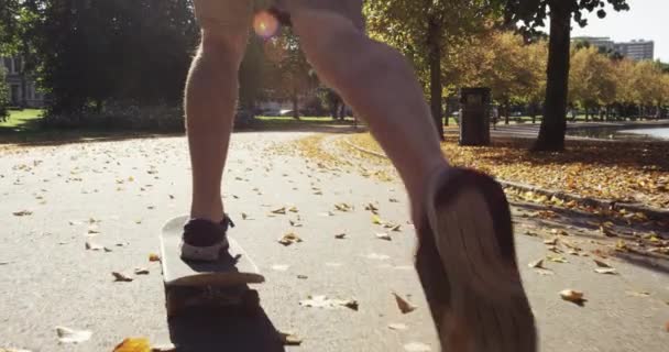 Foot a man skating through a park on his skateboard alone. A man skateboarding on the asphalt through a park on a sunny day. An active man skating through a garden outside — Stock Video