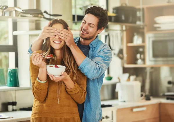 Adivinha quem? Tiro de um jovem afetuoso cobrindo seus olhos namoradas enquanto ela come café da manhã em sua cozinha. — Fotografia de Stock