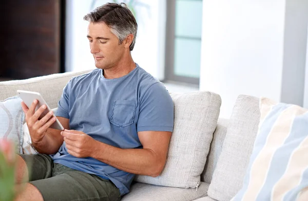 Manter-se atualizado com a tecnologia. Cortado tiro de um homem maduro bonito sentado em seu sofá sala de estar e usando um tablet durante o dia. — Fotografia de Stock