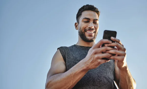 Het opbouwen van spieren met behulp van moderne technologie. Low angle shot van een sportieve jongeman met behulp van een mobiele telefoon tijdens het sporten buiten. — Stockfoto
