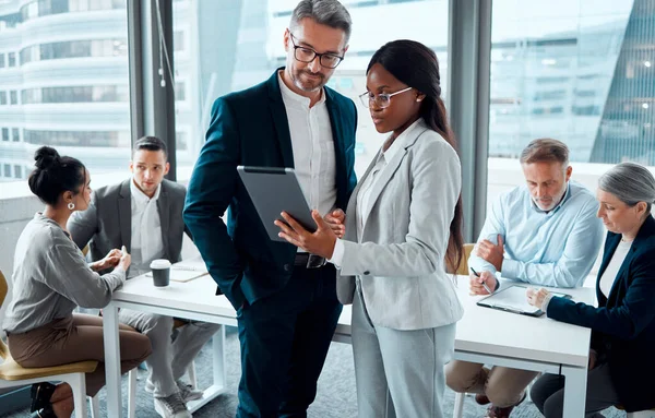 Seja inteligente sobre seus processos de negócios. Tiro de dois empresários usando um tablet digital em um escritório com seus colegas em segundo plano. — Fotografia de Stock