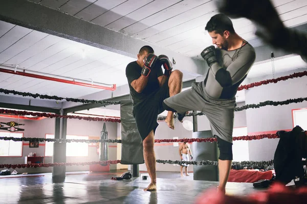 Using some kicks to mix it up. Shot of two young male boxers facing each other in a training sparing match inside of a boxing ring at a gym during the day. — Stock Photo, Image