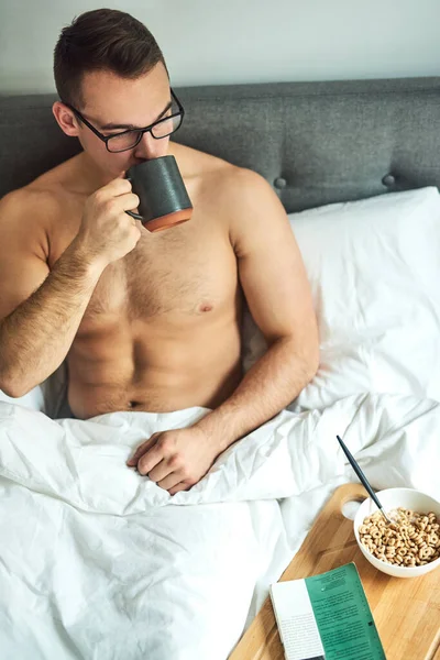 Esta é uma ótima maneira de começar a manhã. Tiro cortado de um belo jovem sem camisa bebendo café e tomando café da manhã na cama em casa. — Fotografia de Stock