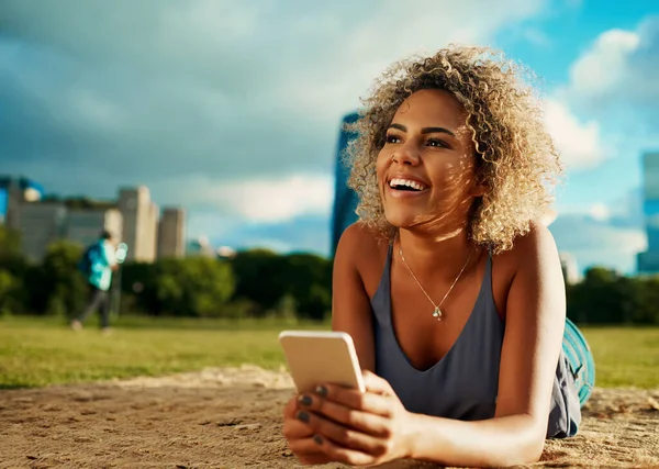 El sol, el parque y el libro electrónico perfecto. Recorte de una atractiva mujer joven usando un teléfono inteligente mientras se acuesta en un parque. — Foto de Stock