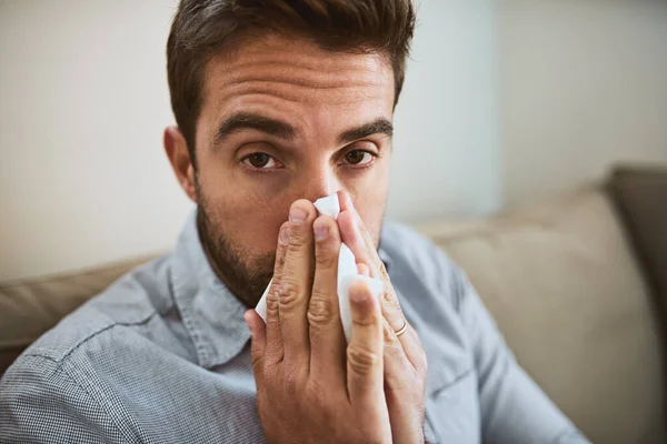 I really dont feel like going to work. Portrait of a uncomfortable looking young man sneezing into a tissue while being seated on a couch at home. — Stock Photo, Image