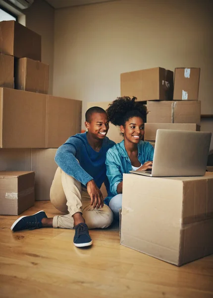 У нас пока нет многого, но мы будем. Съемка веселой молодой пары, просматривающей ноутбук вместе, будучи окруженной картонными коробками внутри дома. — стоковое фото