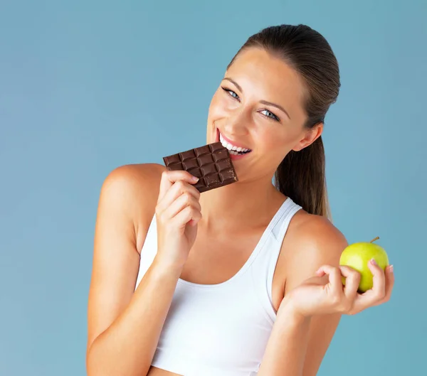 Studio shot of a fit young woman holding an apple while taking a bite of chocolate against a blue background.