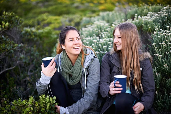 Lachen in de bergen. Shot van twee aantrekkelijke jonge vrouwen genieten van warme dranken tijdens het wandelen. — Stockfoto