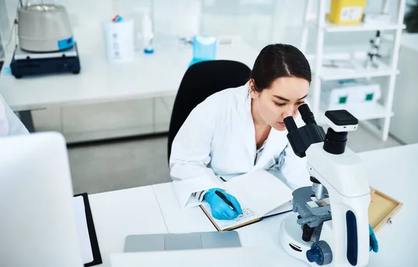 Piszę metodologie badań leków. Zdjęcie młodego naukowca piszącego notatki podczas używania mikroskopu w laboratorium. — Zdjęcie stockowe