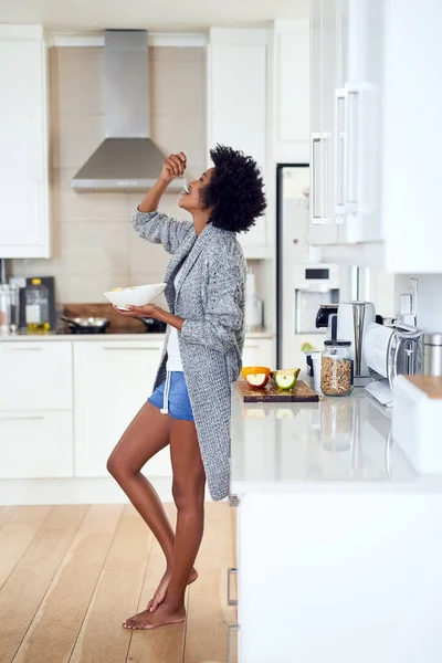 Zaczyna dzień w jej ulubiony sposób. Strzał zrelaksowanej młodej kobiety jedzącej śniadanie stojąc w kuchni w domu. — Zdjęcie stockowe