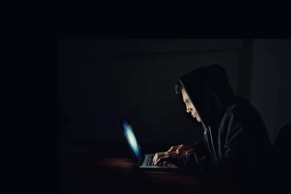 Ayudándose a sí mismo con sus contraseñas. Fotografía de un pirata informático con capucha usando un ordenador portátil en la oscuridad. — Foto de Stock