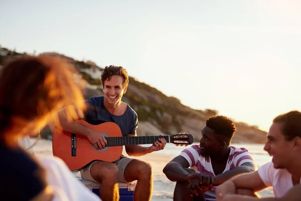 Teman adalah orang-orang yang menginspirasi dan memotivasi kita. Shot of a man playing the guitar while sitting on the beach with his friends. — Stok Foto