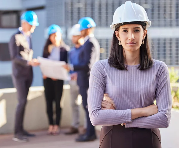 Renovaties zijn aan de gang. Schot van een jonge zakenvrouw met gevouwen armen en een helm op terwijl haar collega 's achter haar staan. — Stockfoto