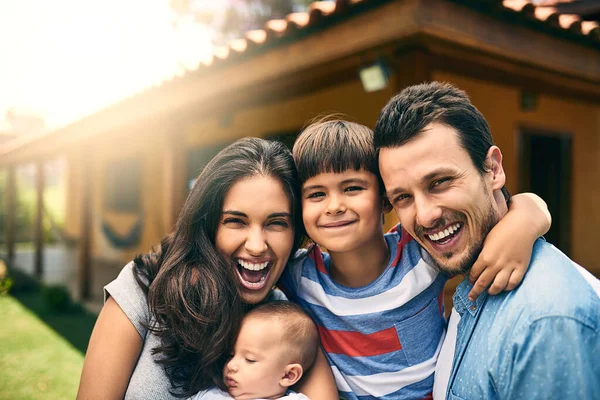 La nostra grande famiglia felice. Ritratto ritagliato di una giovane famiglia felice di quattro persone all'esterno con la loro casa sullo sfondo. — Foto Stock