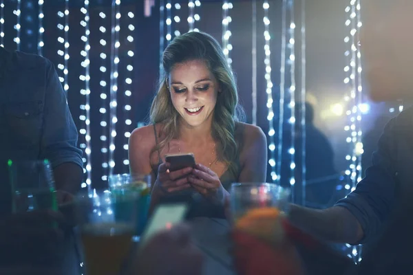 Ven aquí ahora, esta fiesta es increíble. Fotografía de una mujer joven escribiendo en su teléfono inteligente mientras estaba sentada en un club nocturno lleno de gente. — Foto de Stock
