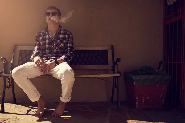 Echando un soplo en el porche. Un disparo de un joven sentado en un banco afuera y fumando un cigarrillo. —  Fotos de Stock