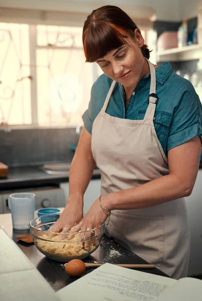 Getting your hands dirty is apart of the recipe. Shot of an attractive young woman reading a recipe from a book while baking at home. — Stock Photo, Image