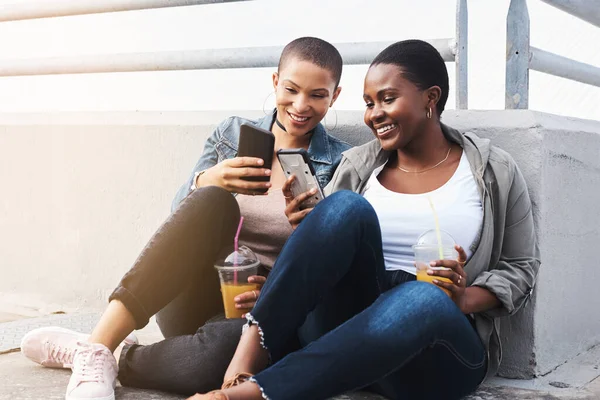 Los verdaderos amigos siempre comparten tendencias. Disparo de dos mujeres jóvenes en la ciudad sentadas mientras reían y sostenían sus bebidas leyendo mensajes de texto. — Foto de Stock