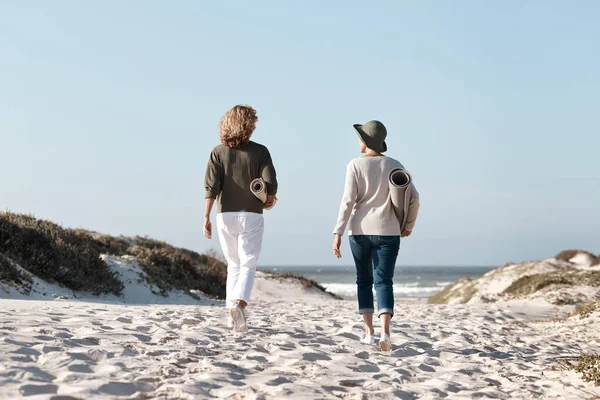 Drar ut for å finne det perfekte stedet. Bilder fra baksiden av to ugjenkjennelige kvinner som går med matta si på stranda. – stockfoto