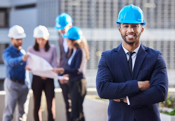 Ik heb het team dat het kan laten gebeuren. Schot van een jonge zakenman met gevouwen armen en een helm op terwijl zijn collega 's achter hem staan. — Stockfoto