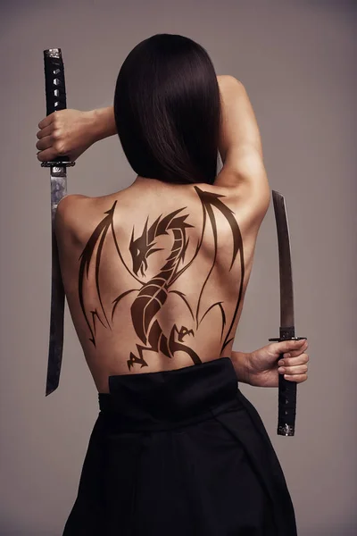 竜の入れ墨の少女。背中に龍の入れ墨で2つの刃を持つ若い女性の武道家の概念的なショット. — ストック写真