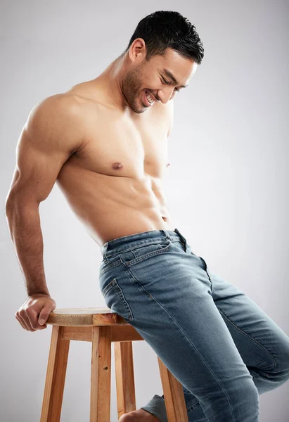 Смеясь над своей дорогой к хорошей жизни. Студийный снимок красивого молодого человека, демонстрирующего свое мускулистое тело, сидящего на стуле на сером фоне. — стоковое фото