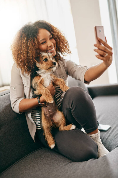 Smile for the camera my darling. Shot of a cheerful young woman taking a selfie with her cute little puppy while being seated on a couch at home.