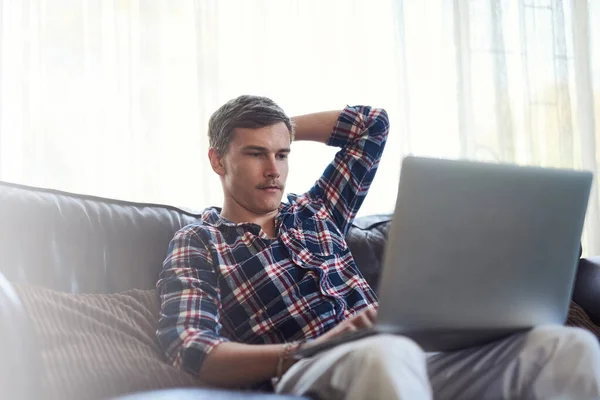 Löytää mielenkiintoista sisältöä verkossa. Ammuttu komea nuori mies käyttää hänen laptop istuessaan sohvalla kotona. — kuvapankkivalokuva