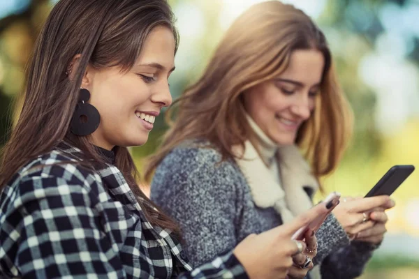 Verbonden blijven, zelfs buiten. Gesneden foto van twee aantrekkelijke jonge vrouwen met behulp van hun mobiele telefoons tijdens het zitten in het park. — Stockfoto