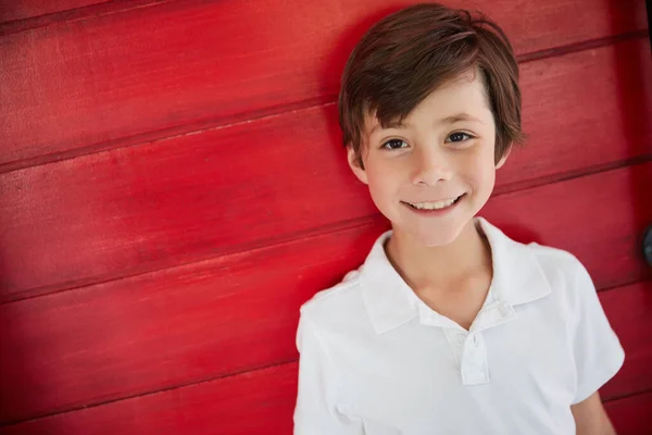 Han är en söt liten kille. Porträtt av en leende liten pojke som står mot en röd vägg. — Stockfoto