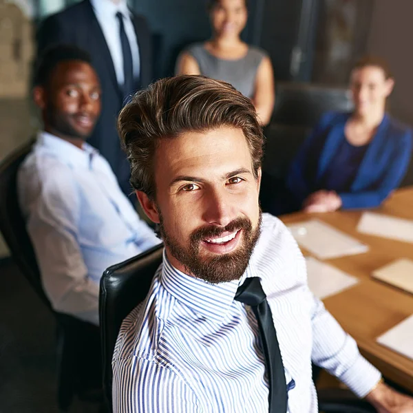 Úspěch svítí na můj tým. Portrét usměvavého podnikatele sedícího v kanceláři s kolegy v pozadí. — Stock fotografie