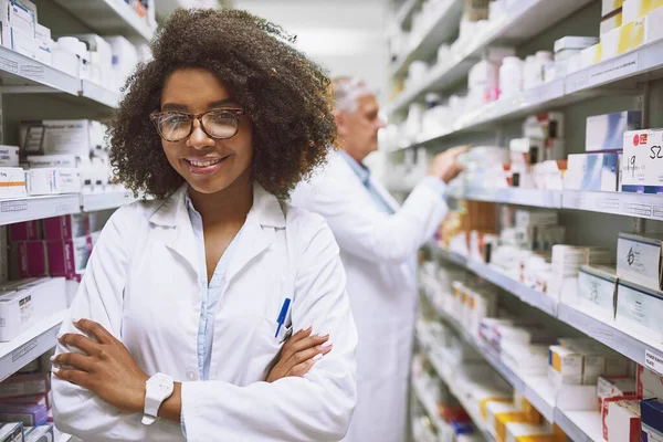 У нас є те, що вам потрібно тут. Портрет веселого молодого жіночого фармацевта, що стоїть зі складеними руками, дивлячись на камеру в аптеці . — стокове фото