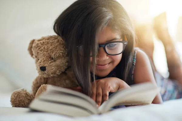 Ми обожнюємо історію в будь-який час. Знімок милої маленької дівчинки, що читає книгу у своїй спальні з плюшевим ведмедем біля неї . — стокове фото
