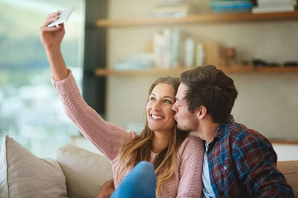Ukazují svou lásku světu. Snímek šťastného mladého páru s selfie s chytrým telefonem, zatímco sedí na gauči doma. — Stock fotografie