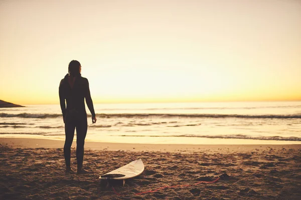 O surf permite-lhe sentir a frescura e a calma da natureza. Foto retrovisada de um jovem surfista olhando para o oceano na praia. — Fotografia de Stock